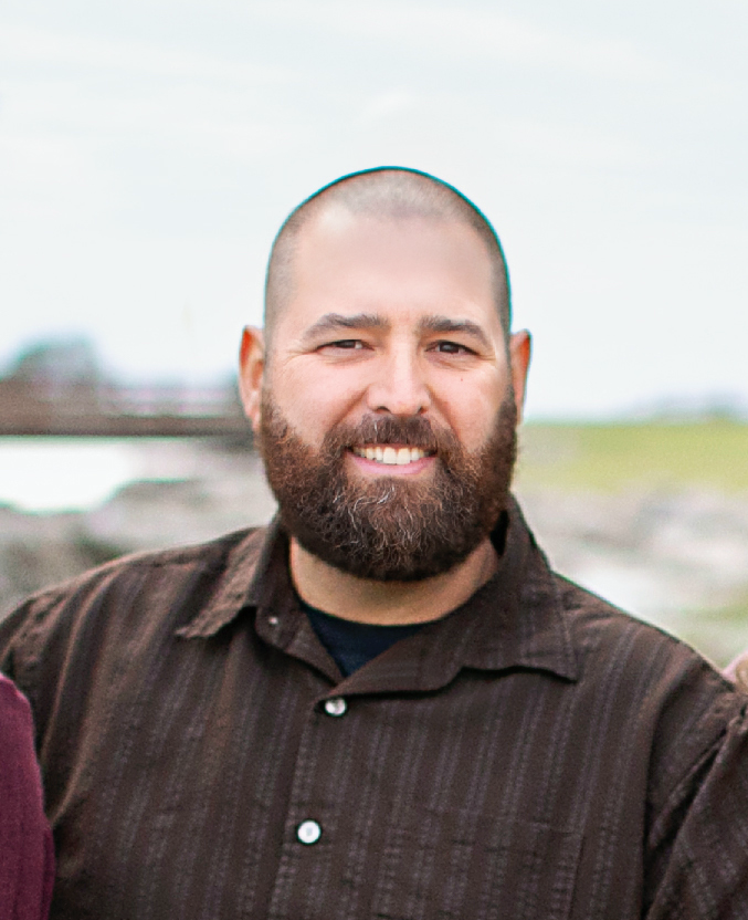 Man with a full brown beard and shaved head smiles, wearing a brown button-down shirt outdoors.