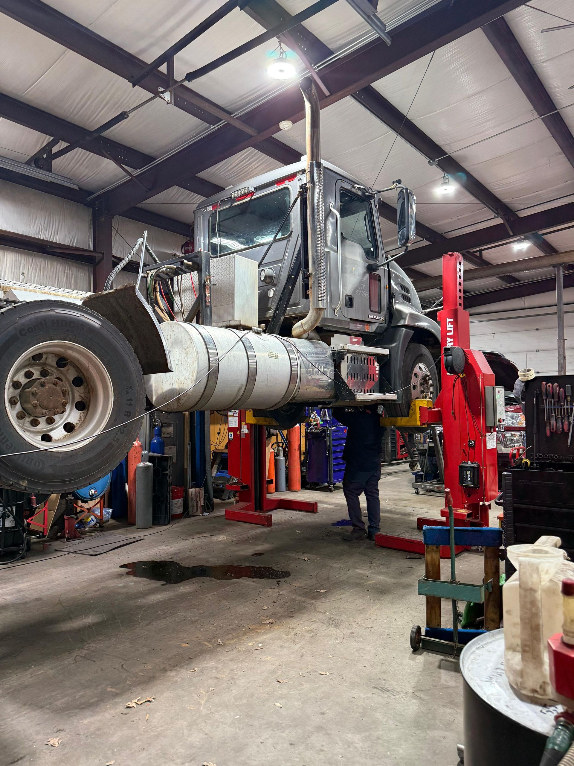 Semi-truck raised on a lift in a repair shop. A person stands beneath. Red and blue equipment is visible.