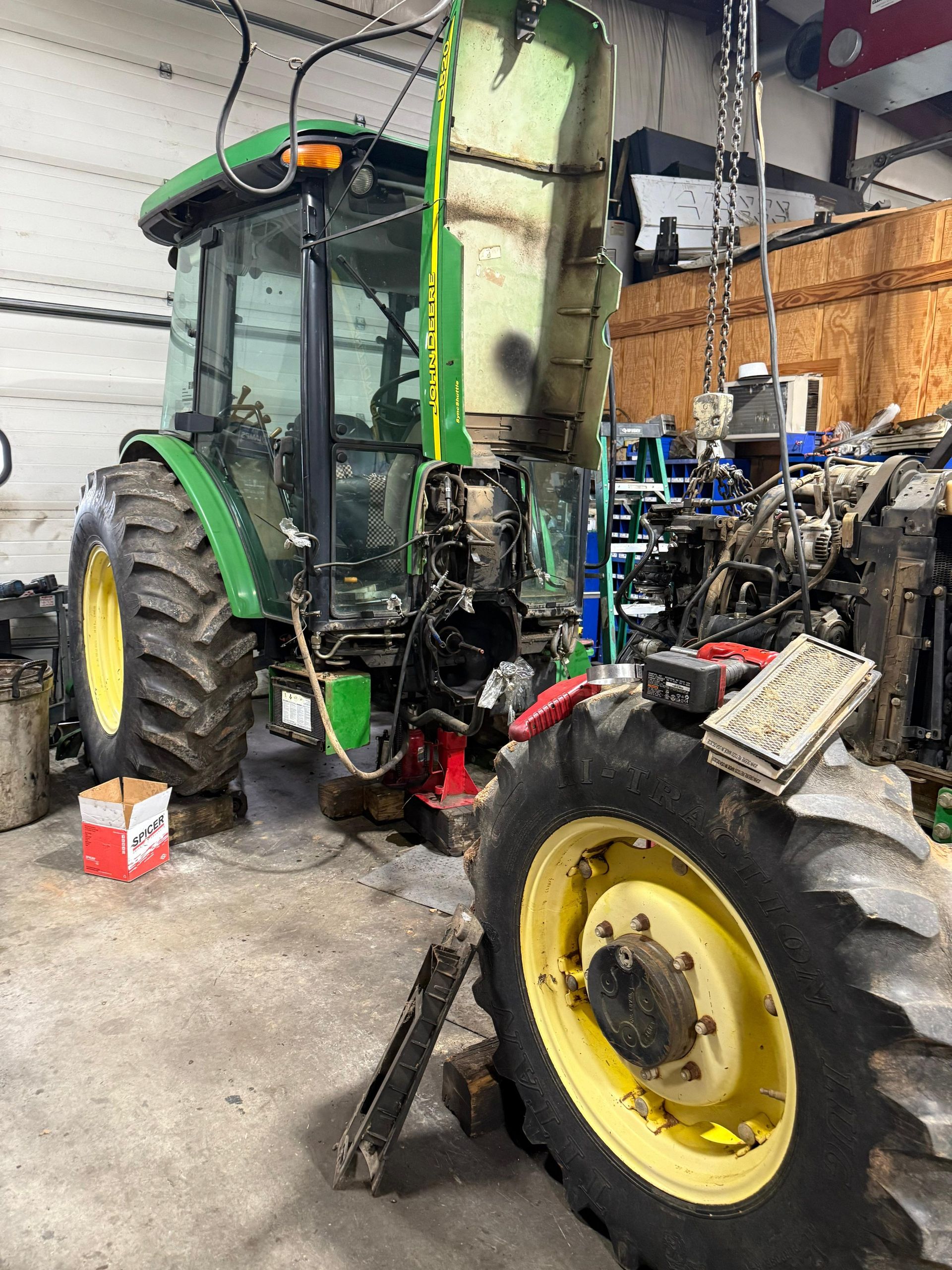 Green John Deere tractor being repaired in a garage, rear tire removed.
