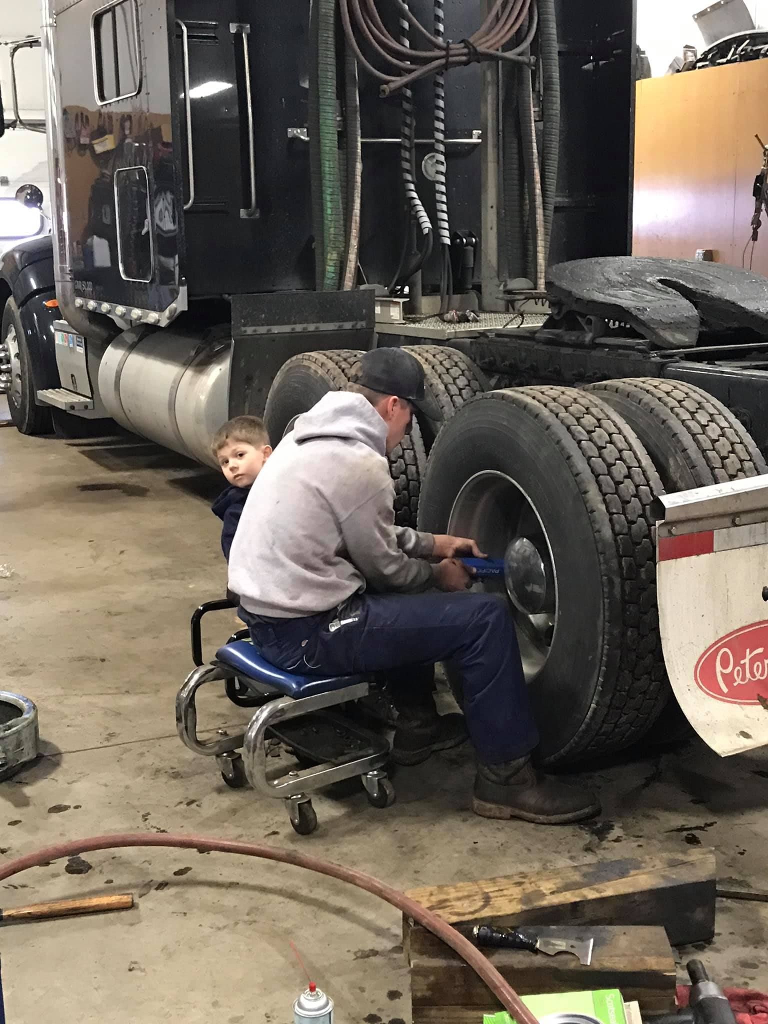 Man in a hoodie working on a truck tire, a child watches nearby in a garage.