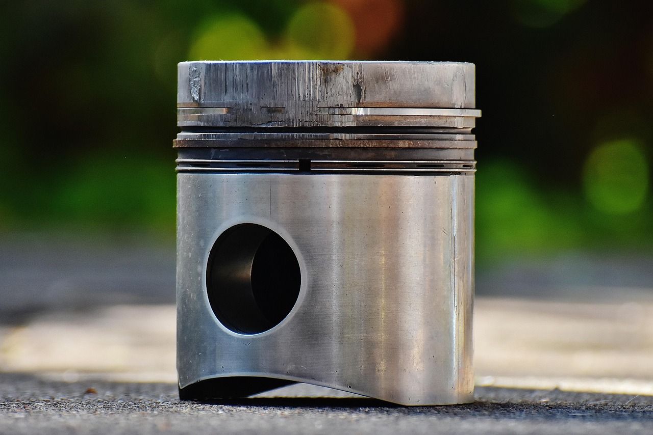 A metal piston with wear marks, a round hole, and ring grooves, set outdoors on a gray surface with blurred green background.