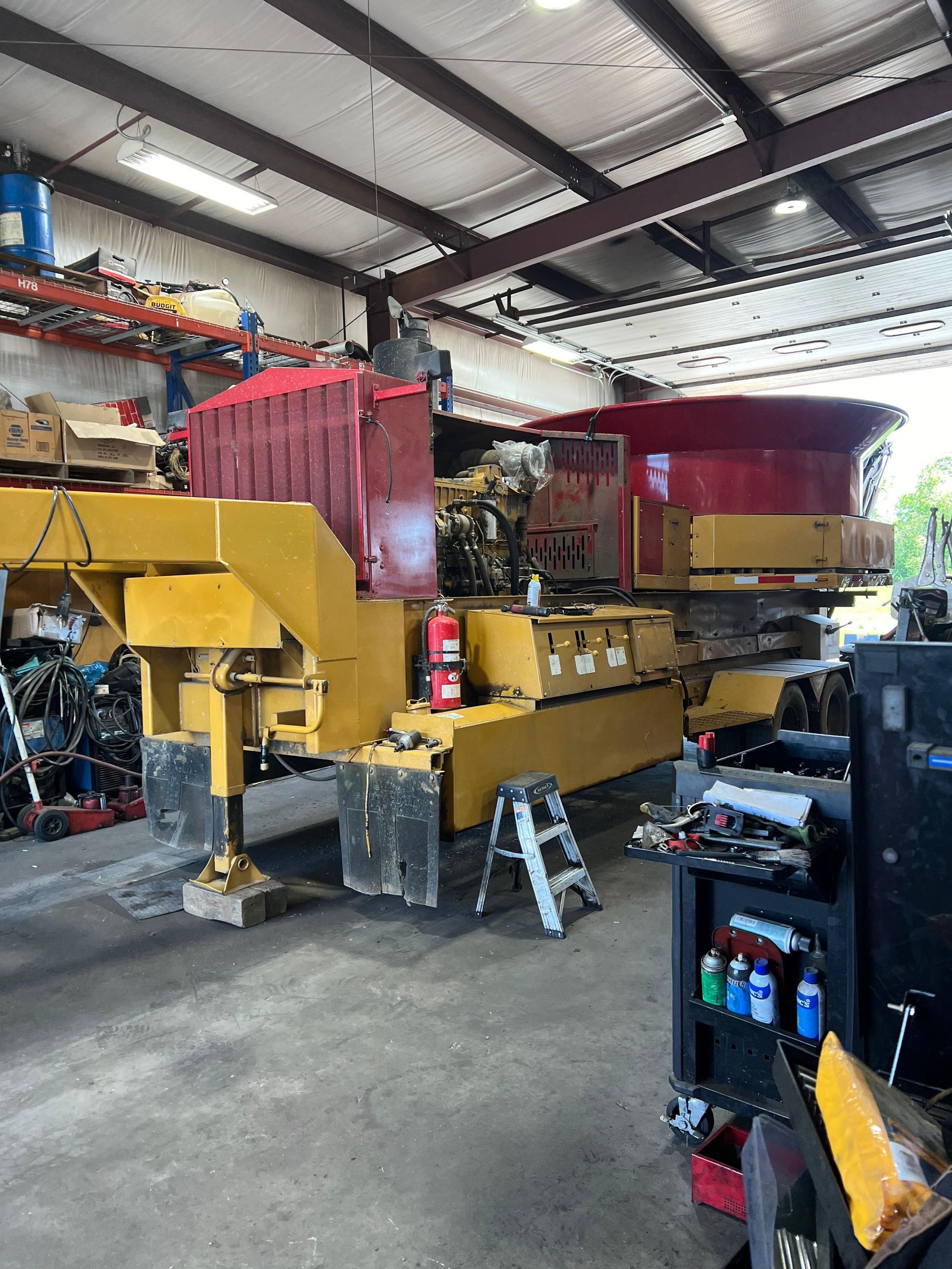 Yellow and red construction equipment inside a repair shop, with tools and a ladder visible.