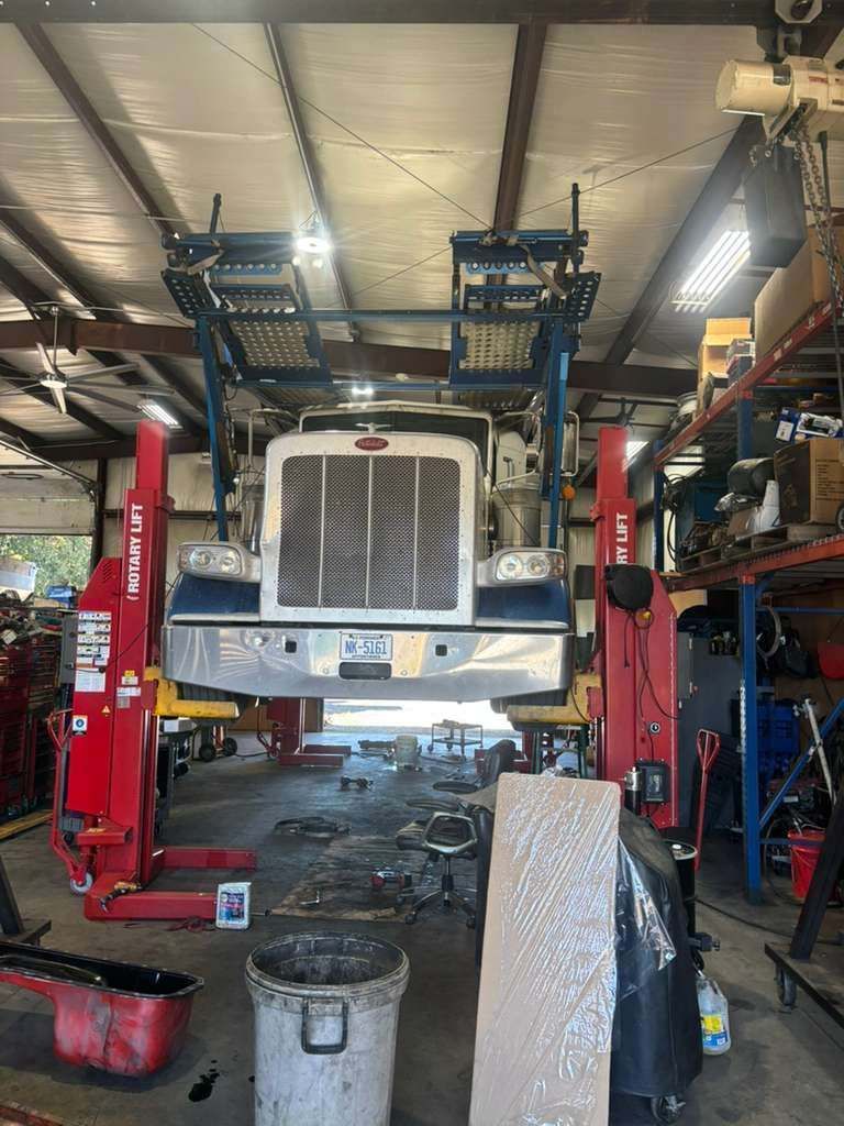 Semi-truck raised on a lift inside a repair shop, with tools and parts scattered on the floor.