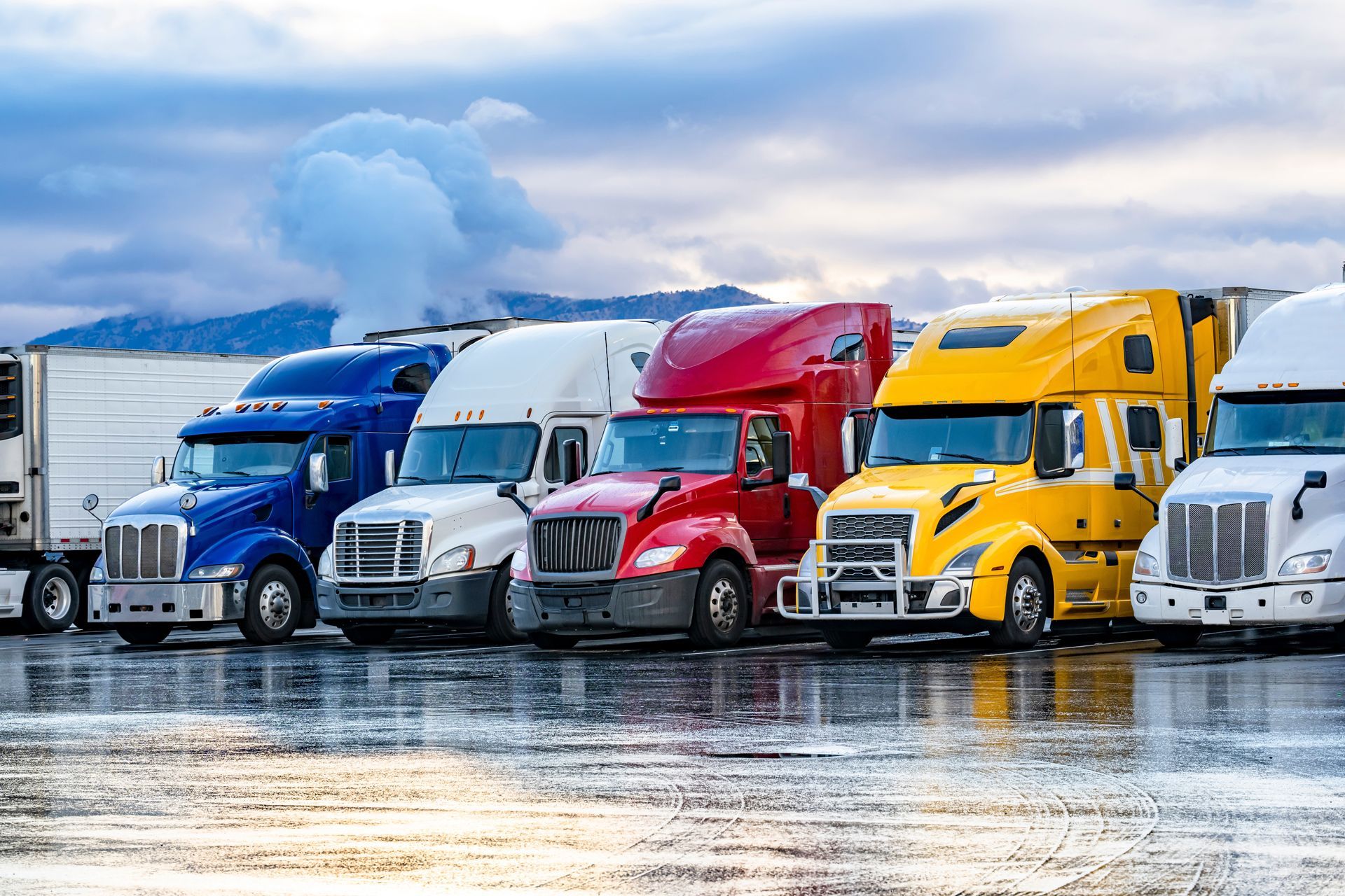 Semi-trucks of various colors parked on a wet lot with mountains in the background.