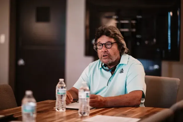 Man in blue shirt, talking, at a table with water bottles, indoors.