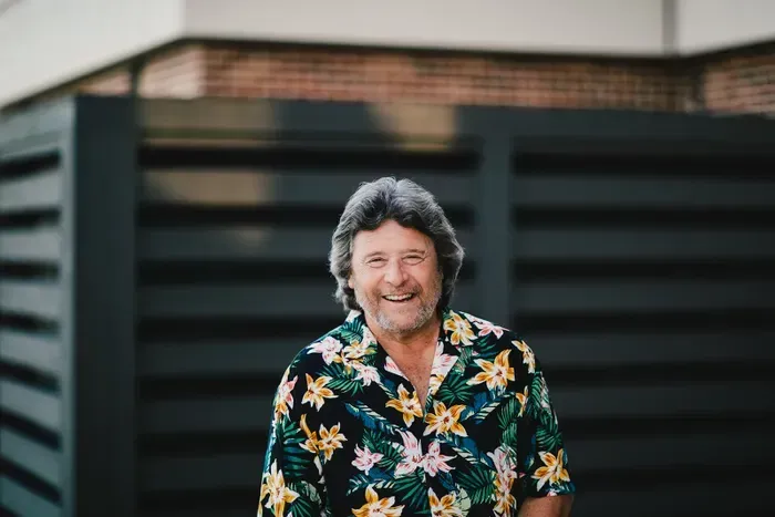 Man smiling, wearing a floral shirt, standing outdoors in front of a dark, slatted fence.