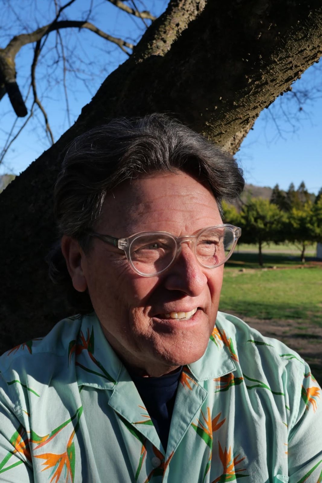 Man smiling outdoors, wearing glasses and patterned shirt, leaning against a tree.