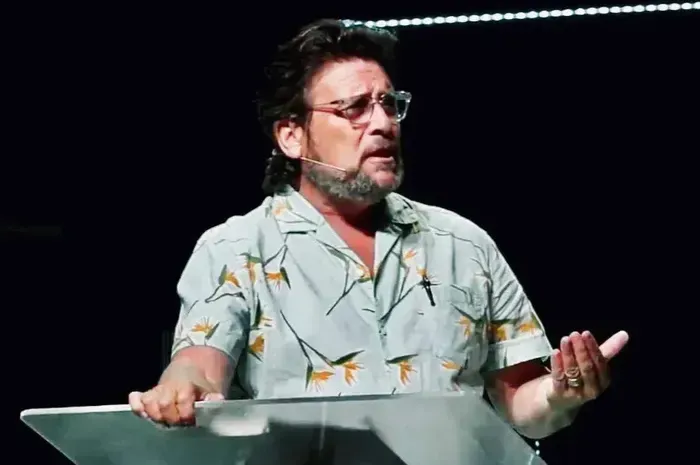 Man speaking at a podium, wearing a patterned shirt and glasses, gesturing with hands. Dark background.