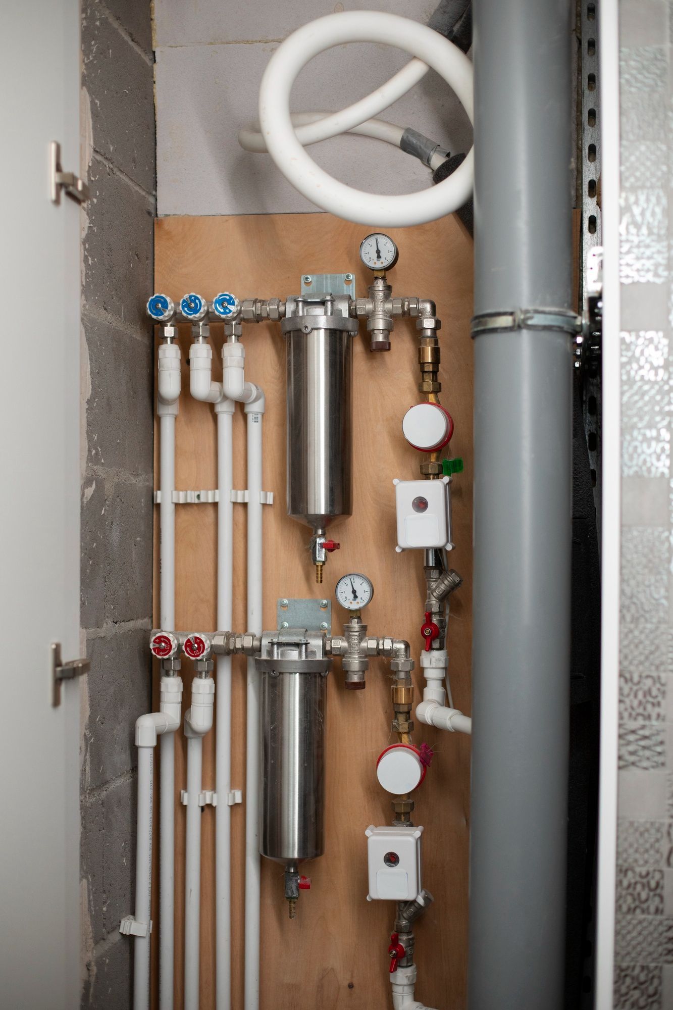 Water filtration system with metal housings, gauges, and white piping mounted on a wooden board next to a gray pipe.