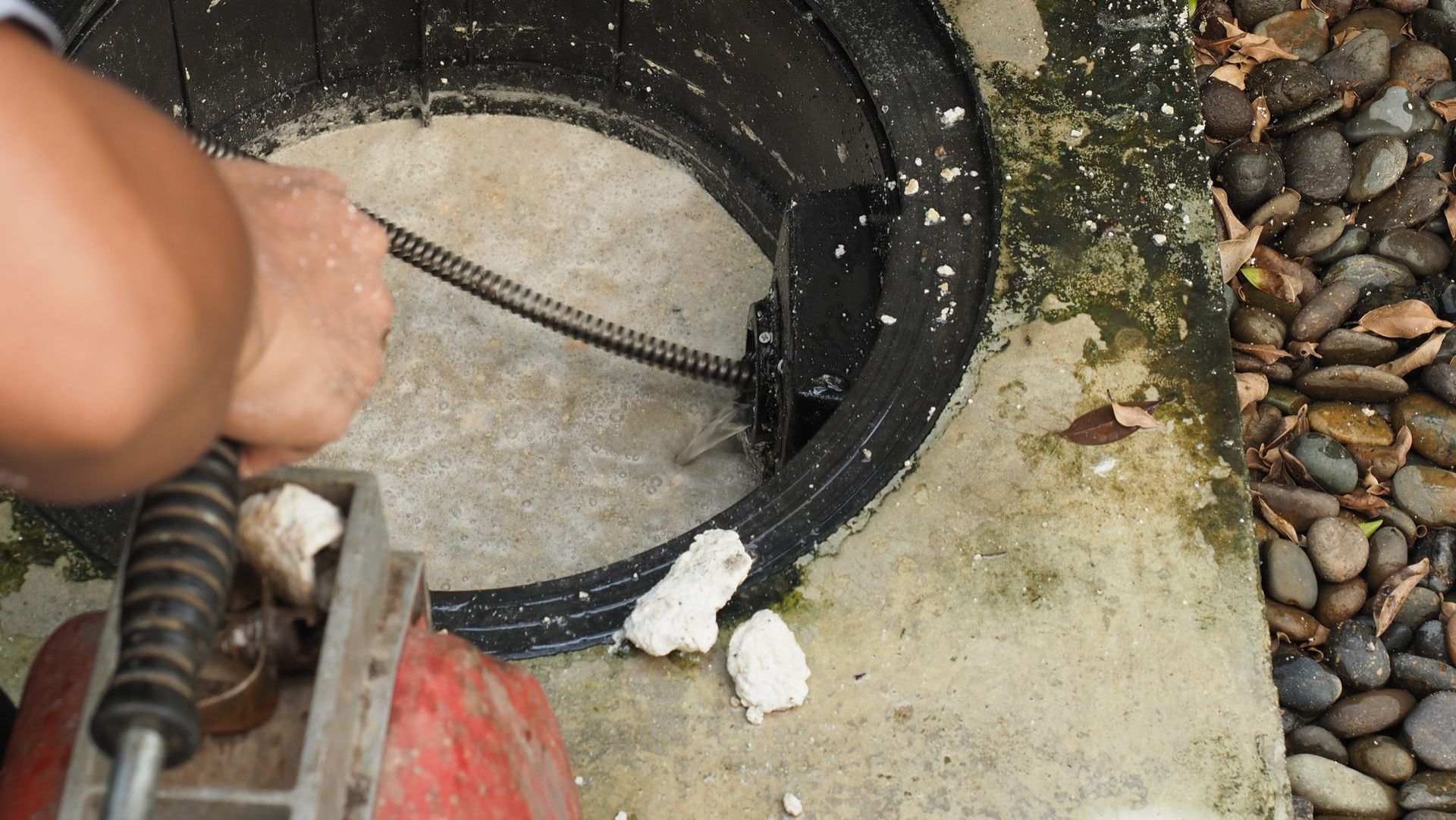 A worker uses a plumbing snake to clear a clog in an outdoor drainage pipe, with gravel visible nearby.