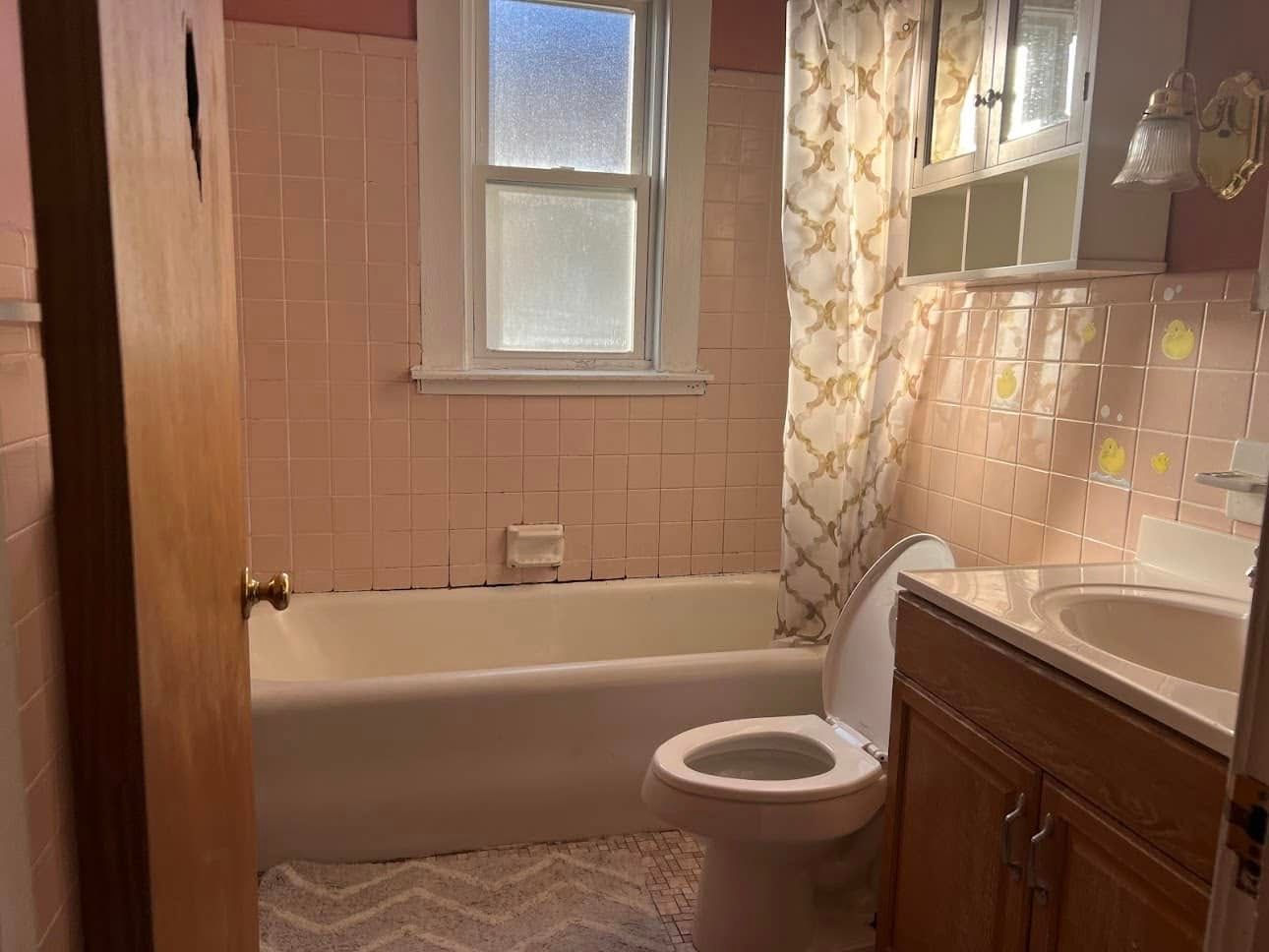 Pink tiled bathroom with a bathtub, toilet, and vanity. A window and patterned shower curtain are visible.