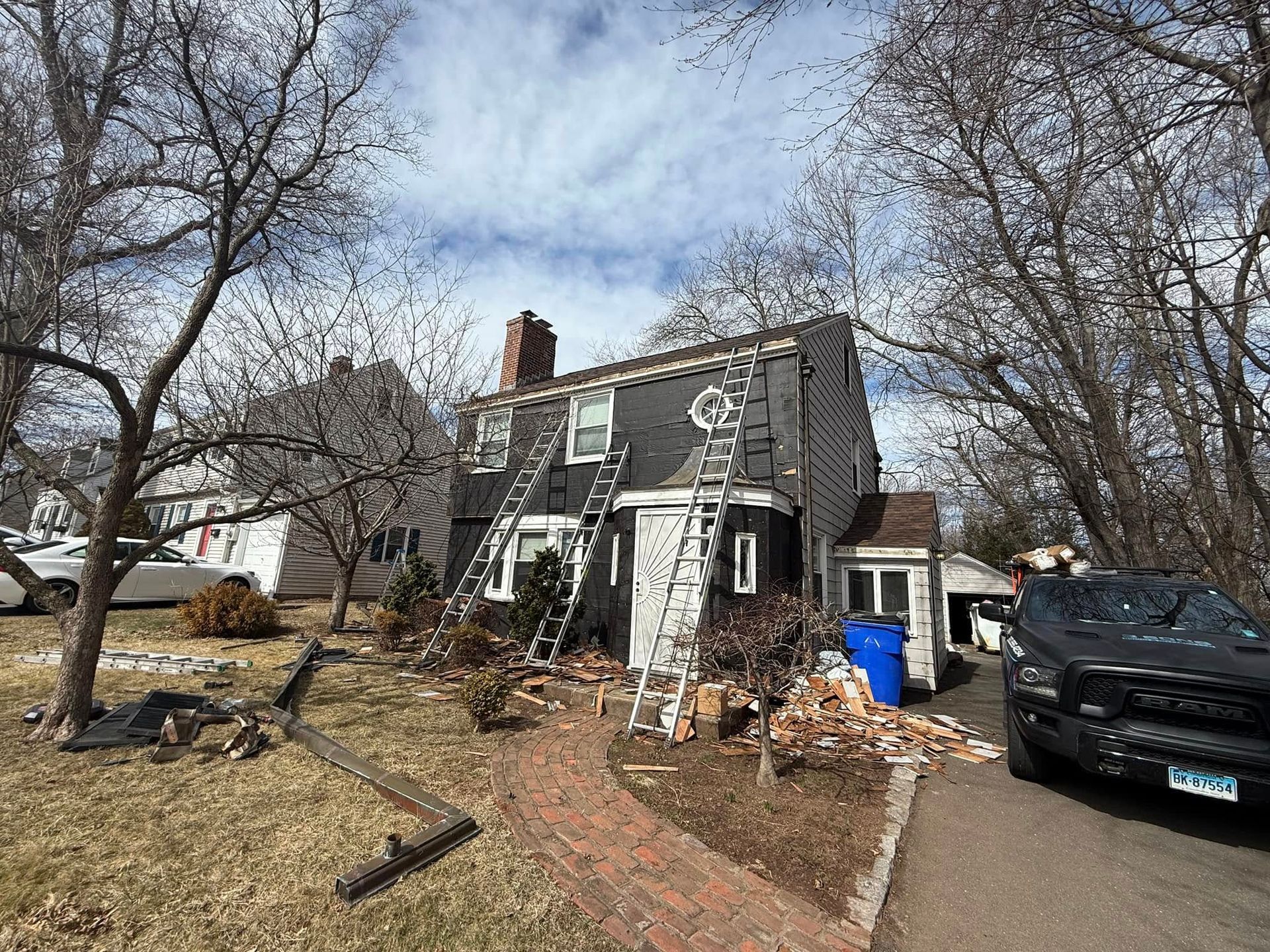 House with ladders, undergoing roof work. Gray siding, brick pathway, and a black truck parked nearby.