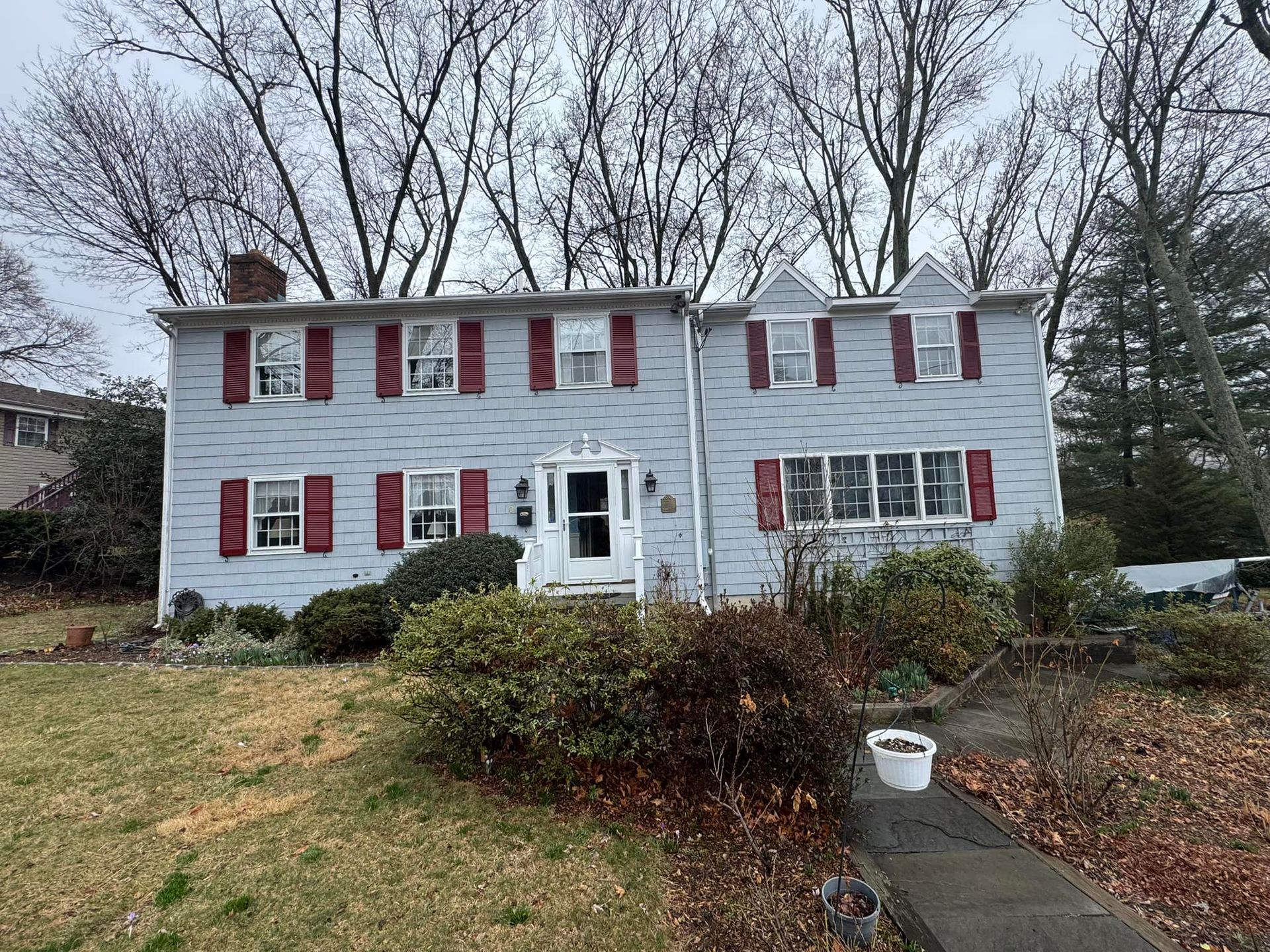 Two-story light blue house with red shutters. Overcast sky, bushes in front, bare trees in the background.