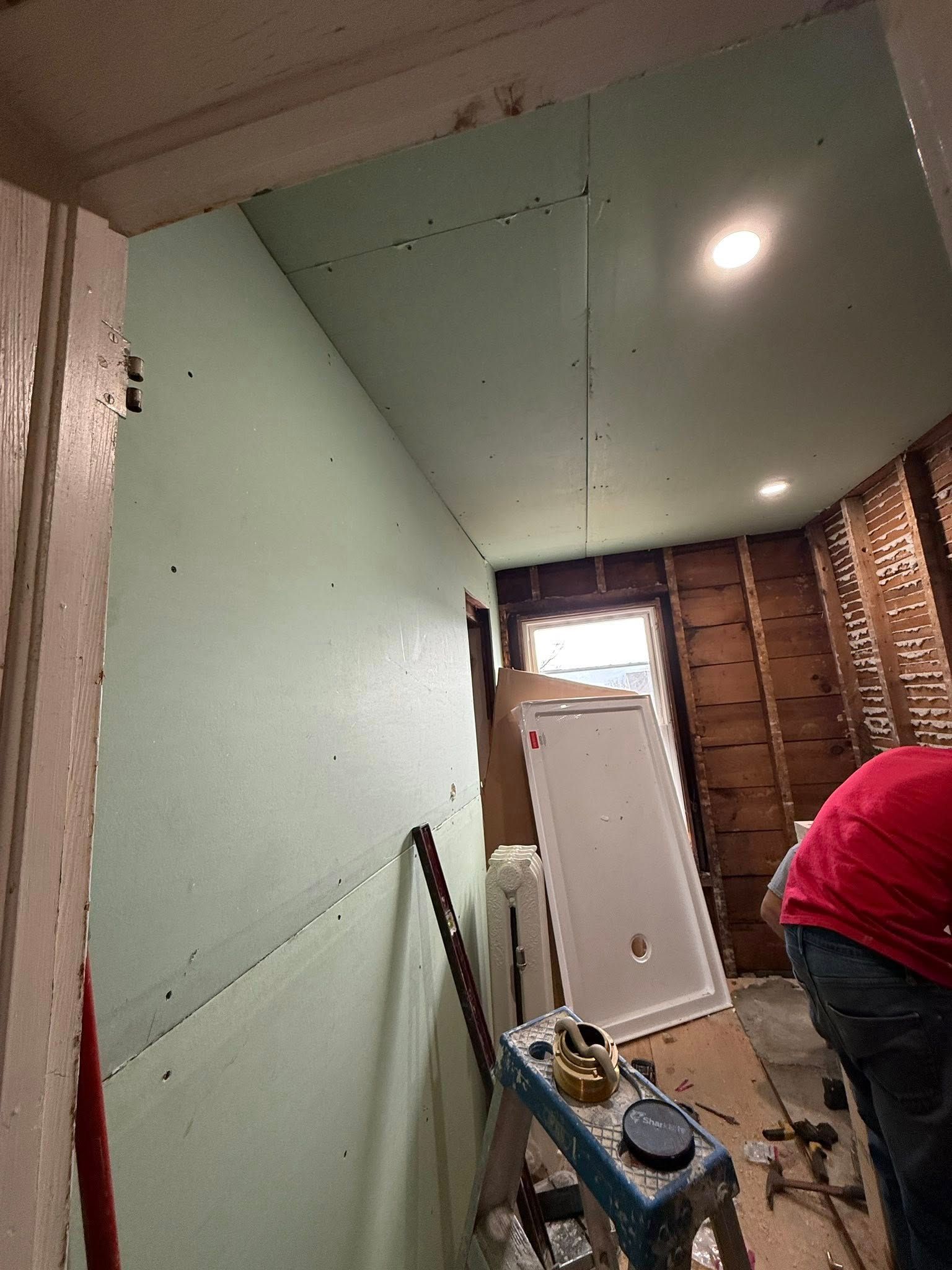 Bathroom under construction, green drywall installed on walls and ceiling. Person in red shirt working.