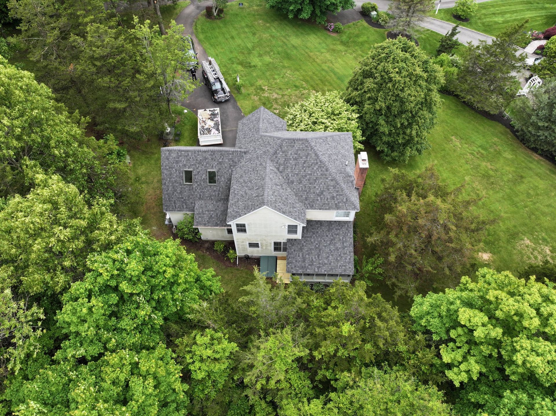 Overhead view of a house with a gray roof, surrounded by green trees and grass, a driveway is visible.