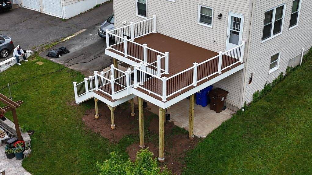 Elevated two-tiered deck on a house with brown decking, white railing, and wooden support beams on a grassy hill.