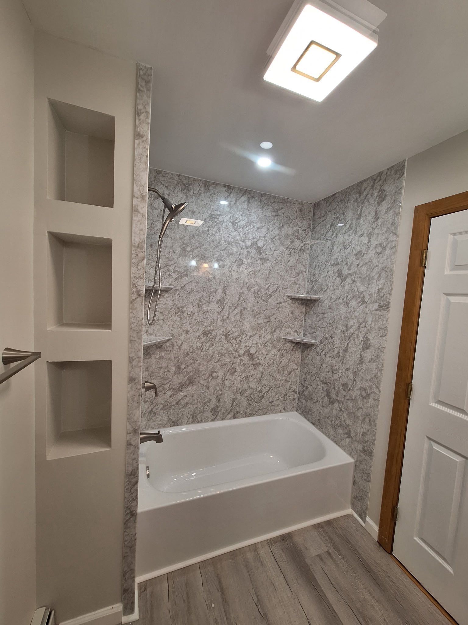 Bathroom with gray and white patterned walls, white bathtub, built-in shelving, and wood-look flooring.