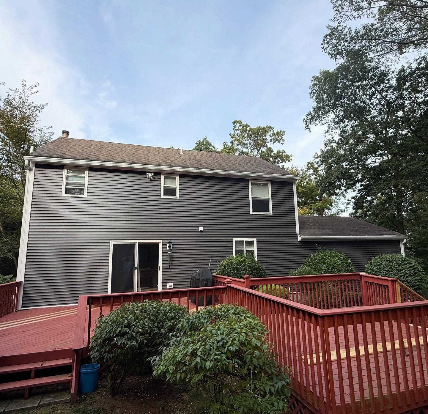 Back of a two-story house with gray siding, red deck, and green shrubs under a cloudy sky.