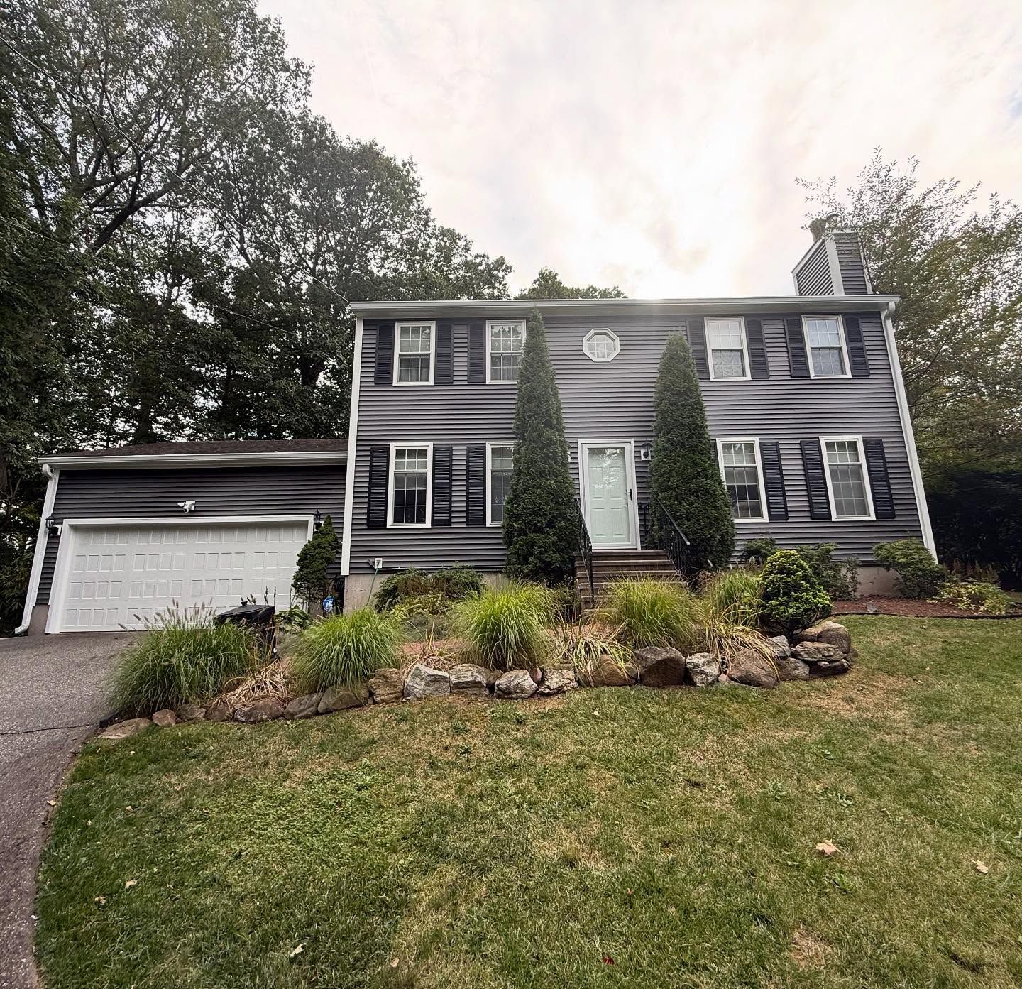 Two-story gray house with white trim, a garage, and a sloped lawn. Trees in the background.