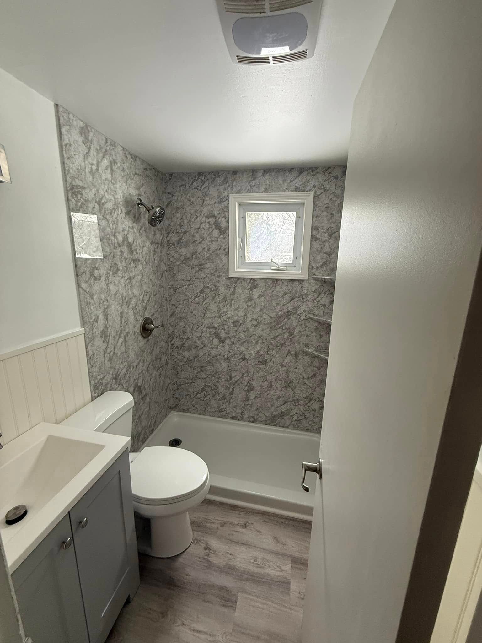 Small bathroom with gray marble-look shower, white toilet, and gray vanity; small window.