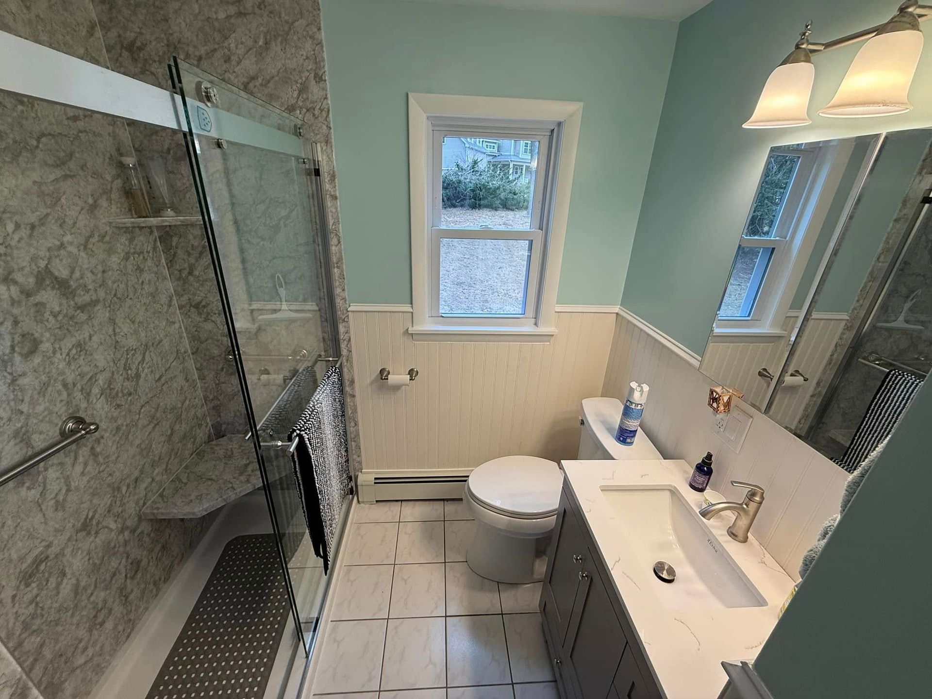 Bathroom with a shower, toilet, and sink; light blue walls, white trim, and a window.