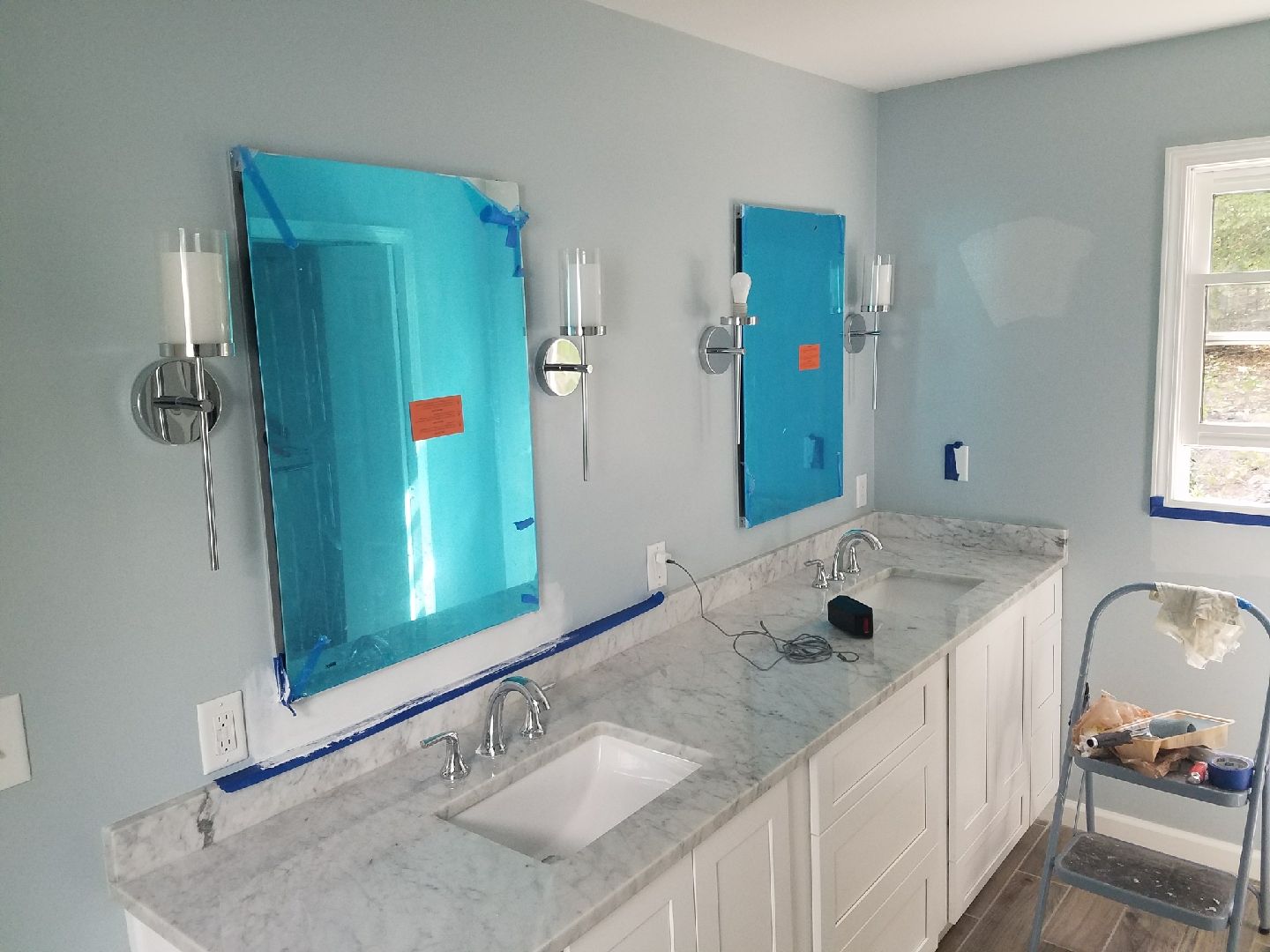 Bathroom with two mirrors covered in protective blue film, double vanity, and wall sconces.