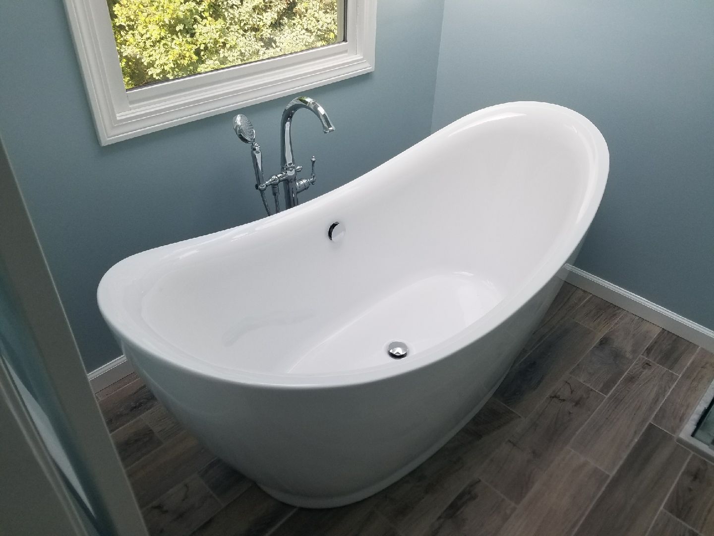 White oval bathtub in a blue-walled bathroom with a window. Chrome faucet. Wood-look tile floor.