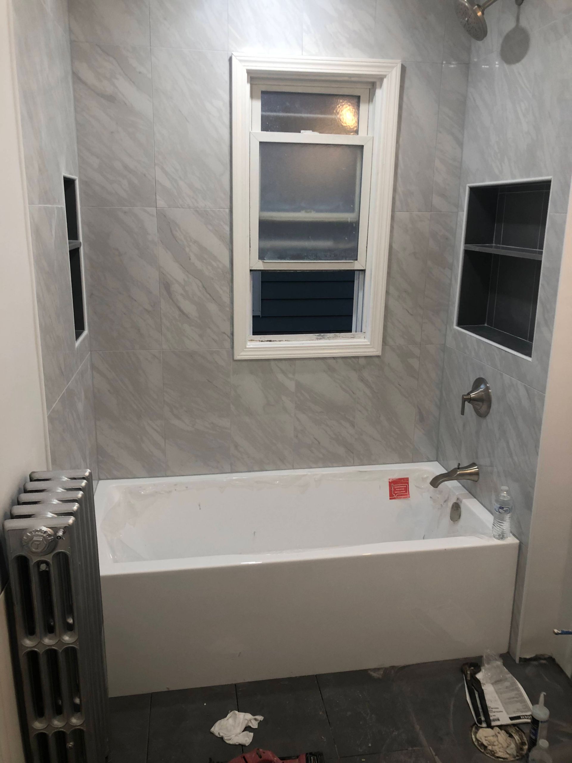 Bathroom renovation: white bathtub, gray marble-look tile, window, built-in shelves, radiator.
