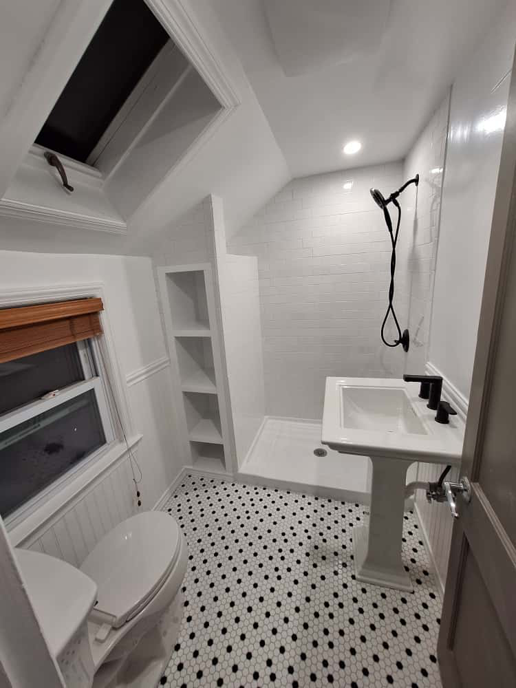 White bathroom with black and white tiled floor, walk-in shower, pedestal sink, and skylight.