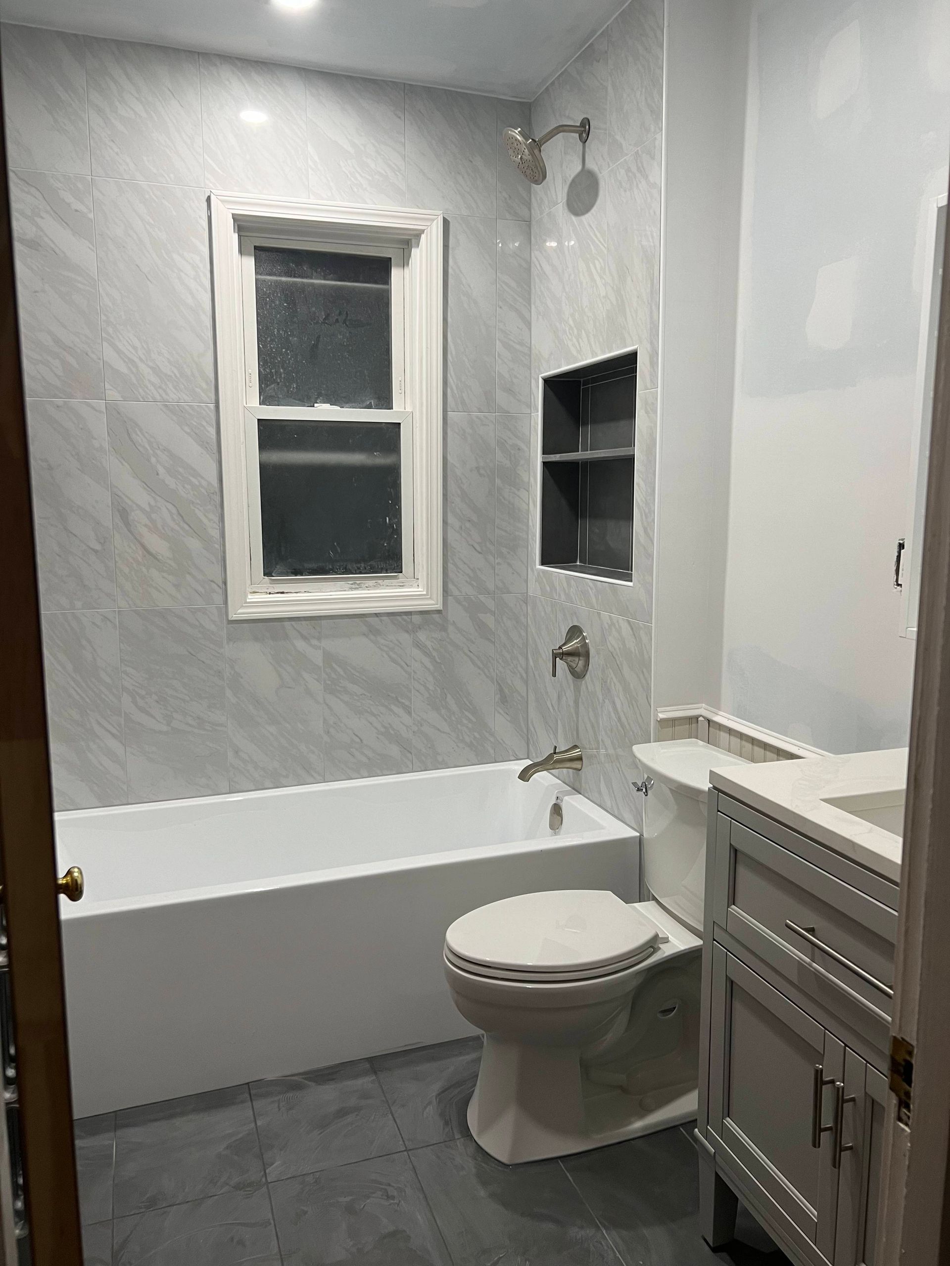 Newly renovated bathroom with white tub, toilet, vanity, and gray tiled walls.