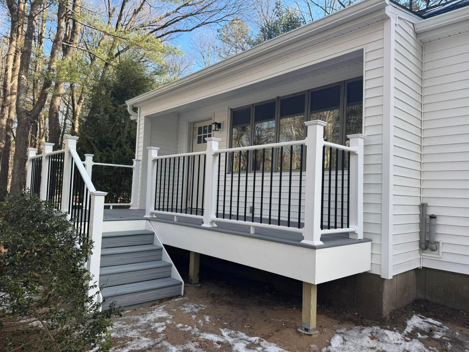 White deck with black railing and steps attached to a white house with windows; set in a yard with trees.