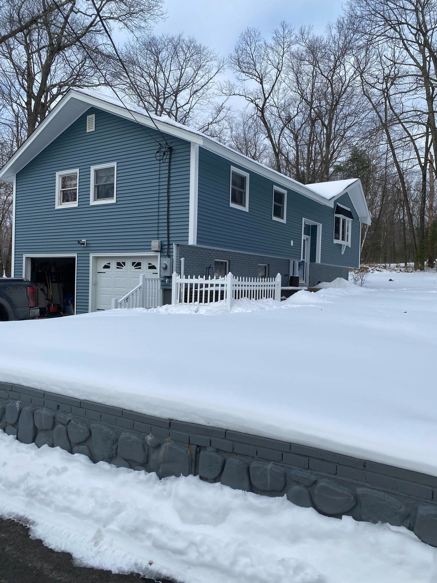 Blue house covered in snow with a white garage door, and a stone wall.