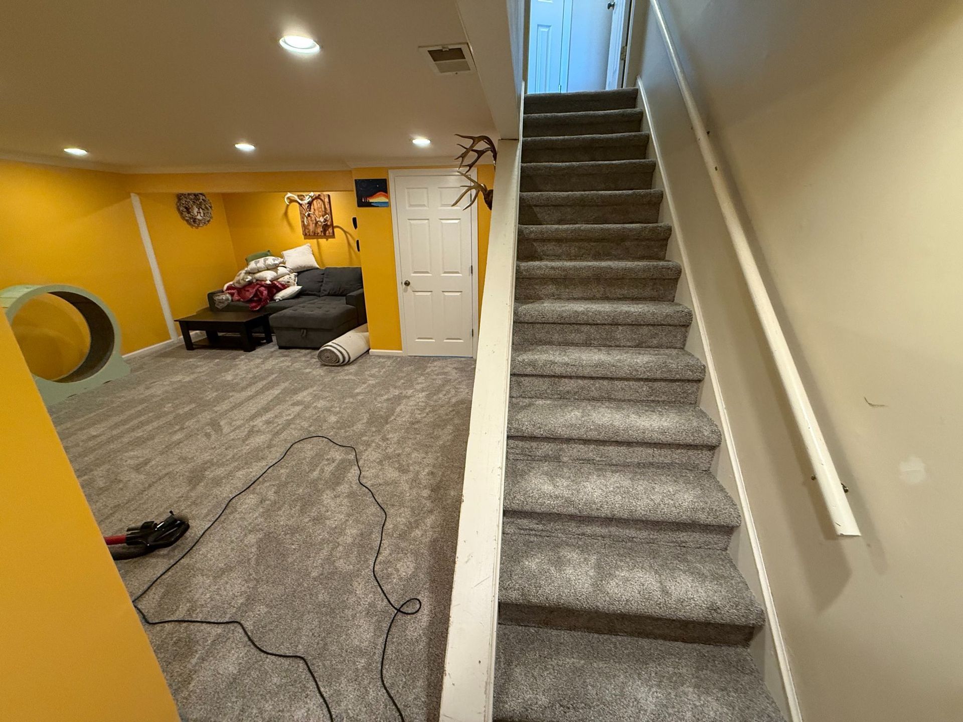 Basement with carpeted stairs, yellow and white walls, seating area, and closed white door.