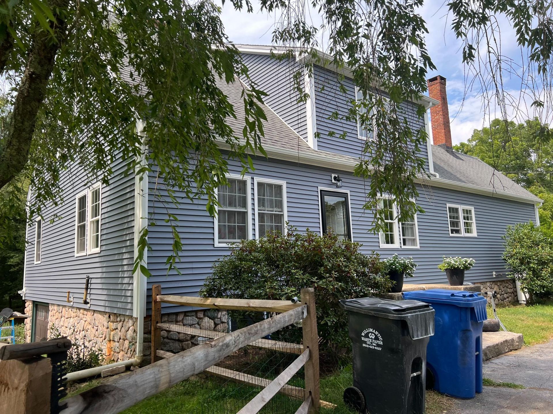Blue clapboard house with stone foundation, wooden fence, and trash bins.