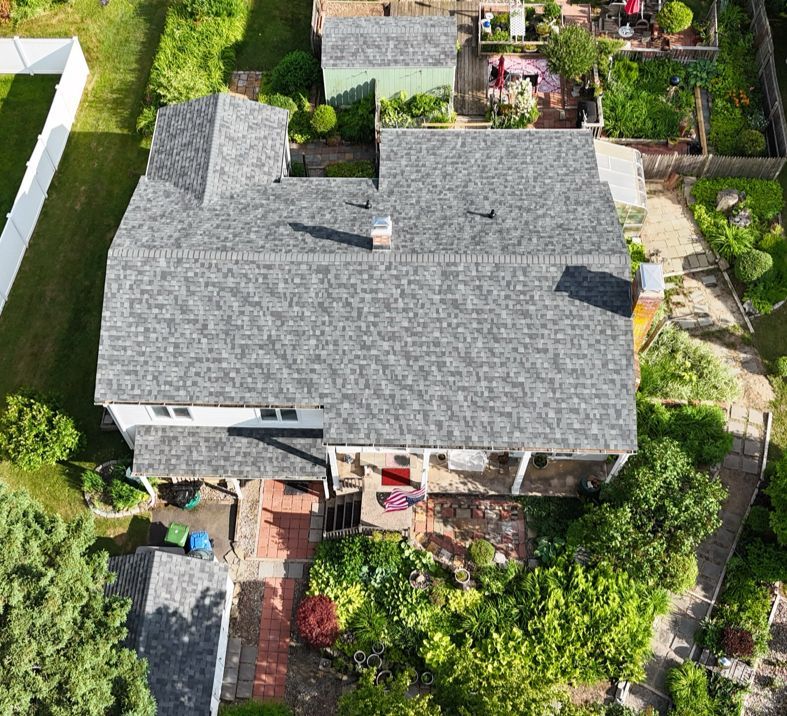 Aerial view of a gray-shingled house with surrounding landscaping, brick patio, and white fence.