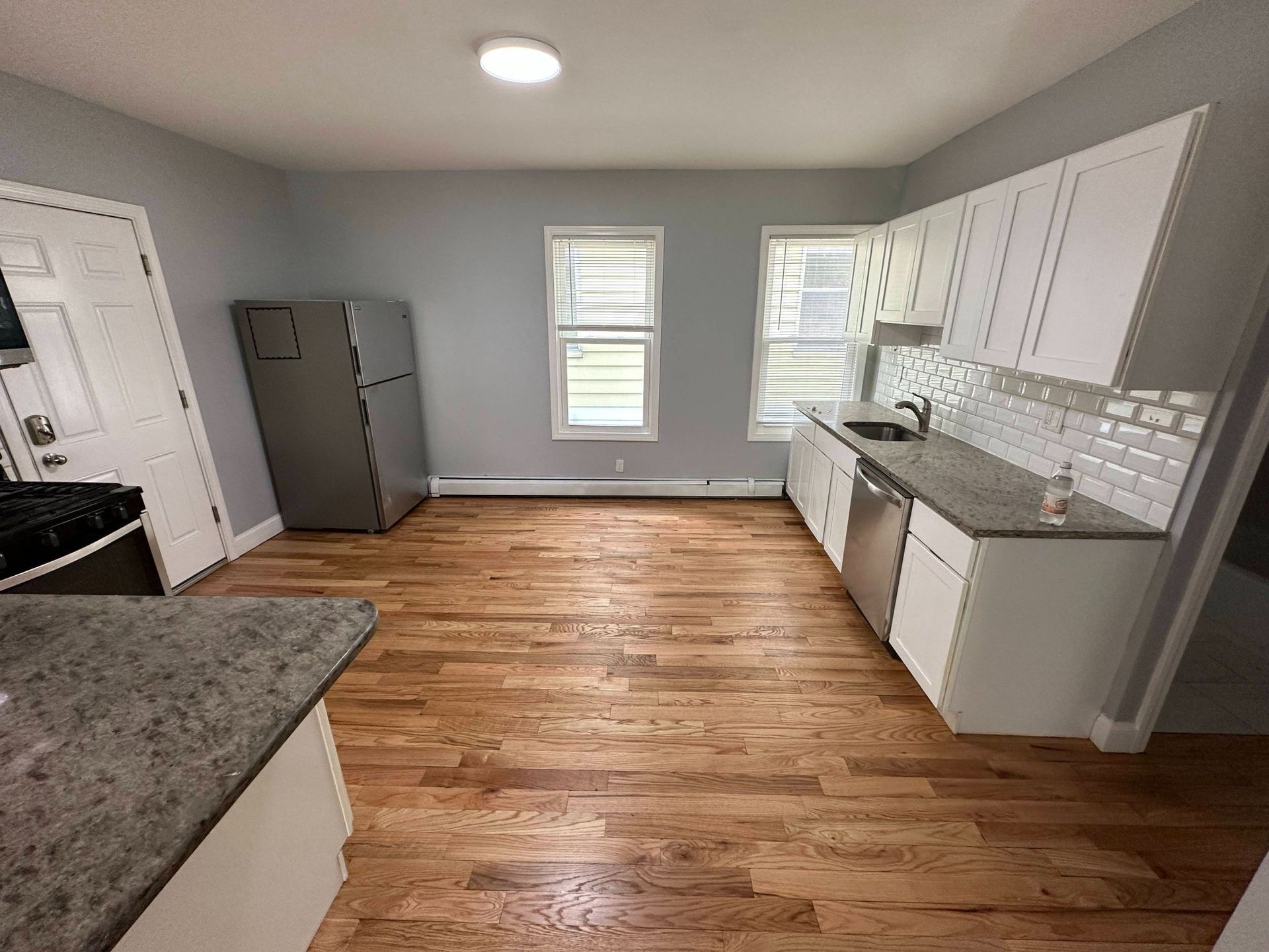 Kitchen with wood floor, white cabinets, gray countertops, stainless steel appliances, and two windows.