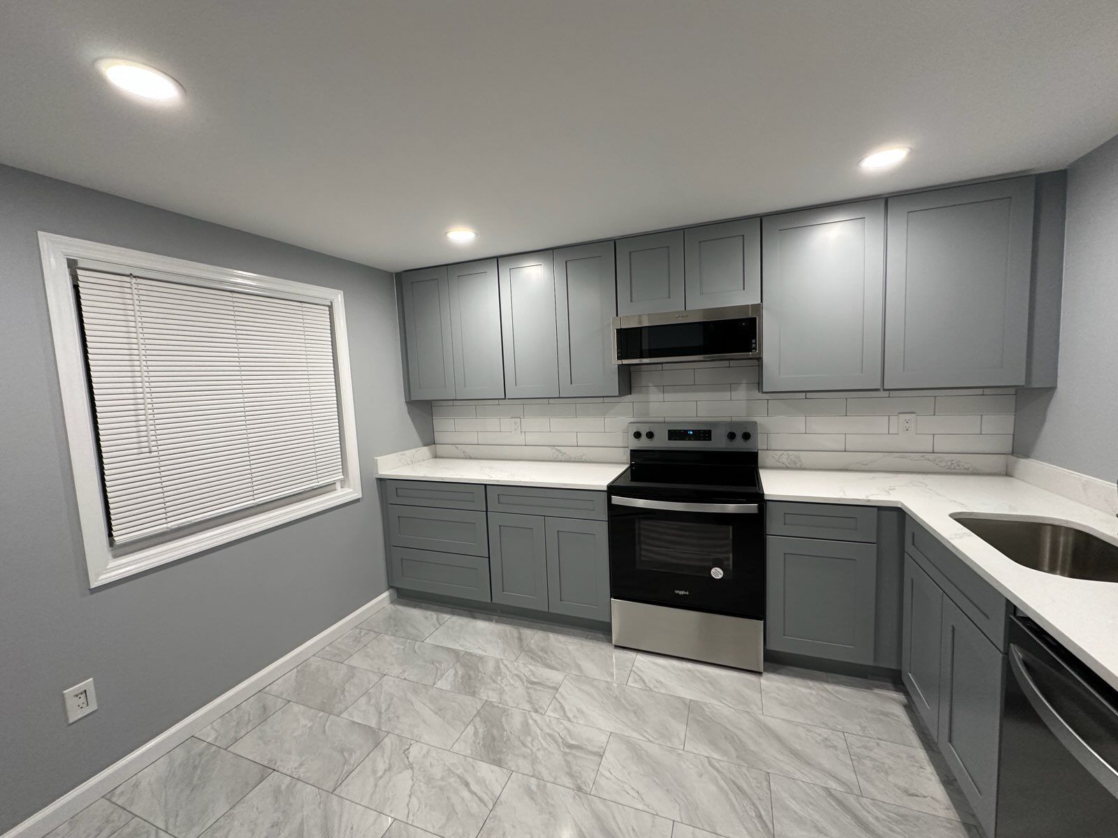 Grey and white kitchen with cabinets, stove, and sink. Window on left.