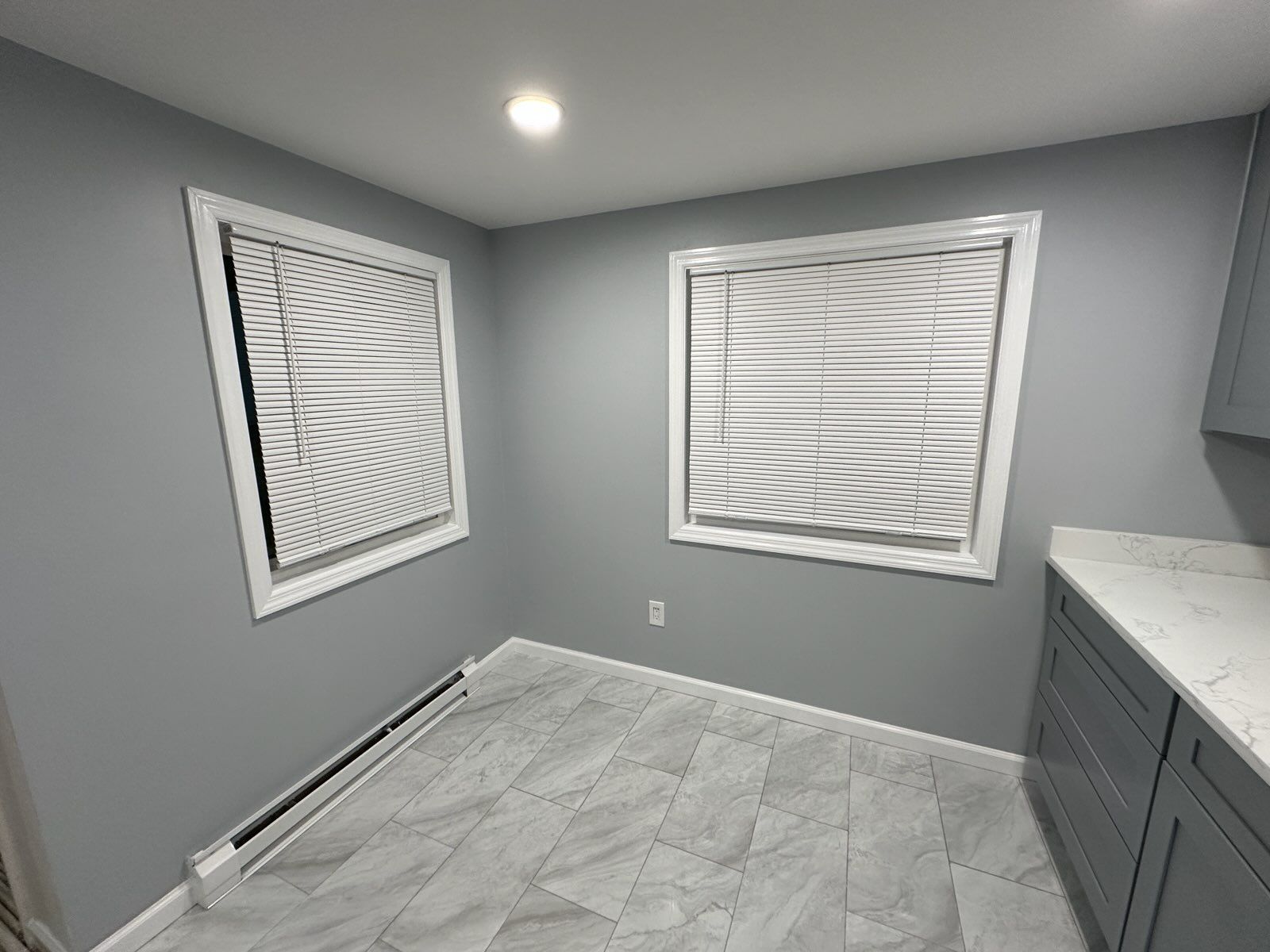 Empty gray kitchen corner with two windows, blinds, and gray cabinets.