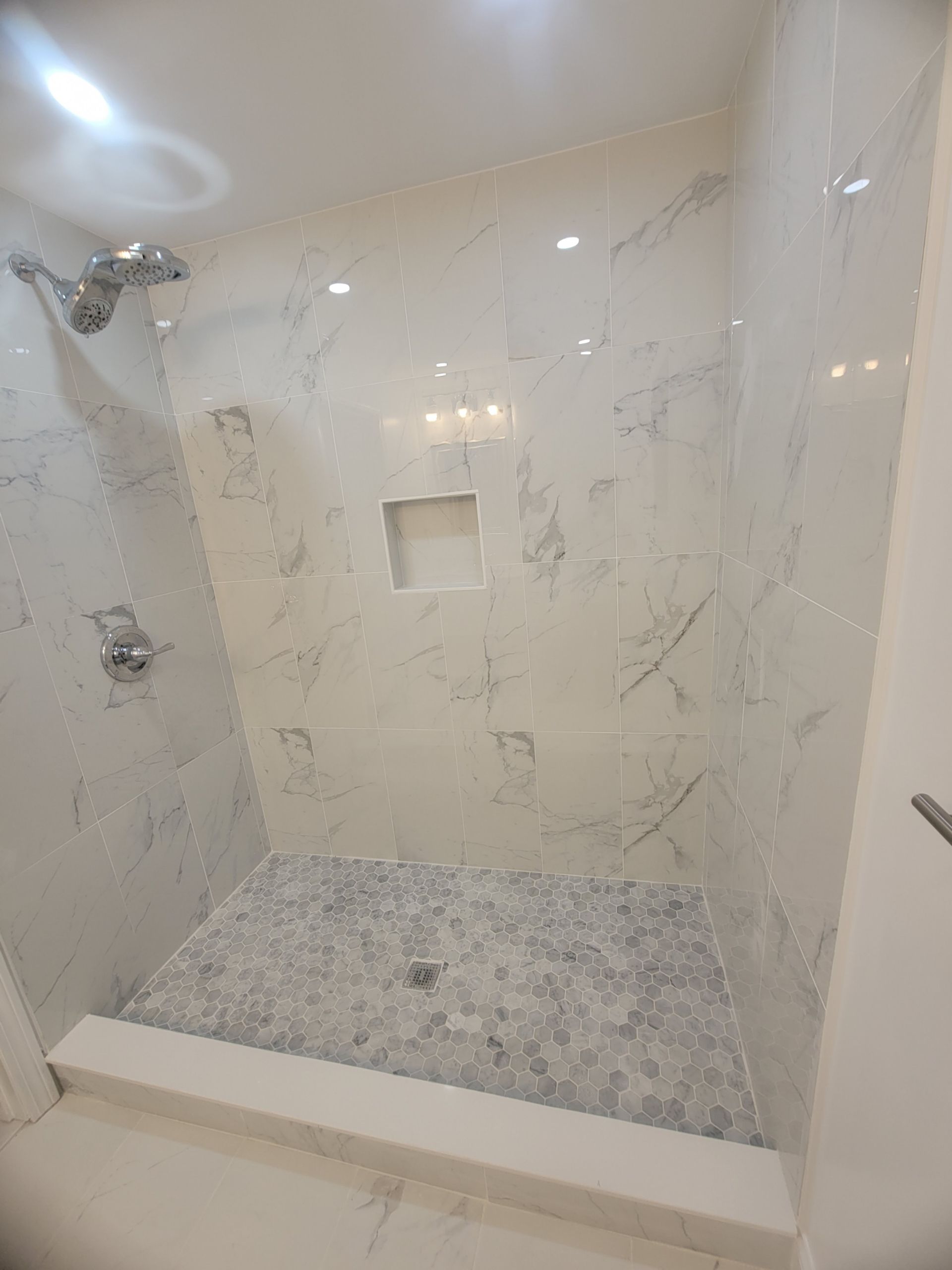 Shower with marble-look tile walls, pebble floor, and recessed shelf. Chrome fixtures, step-up entry.