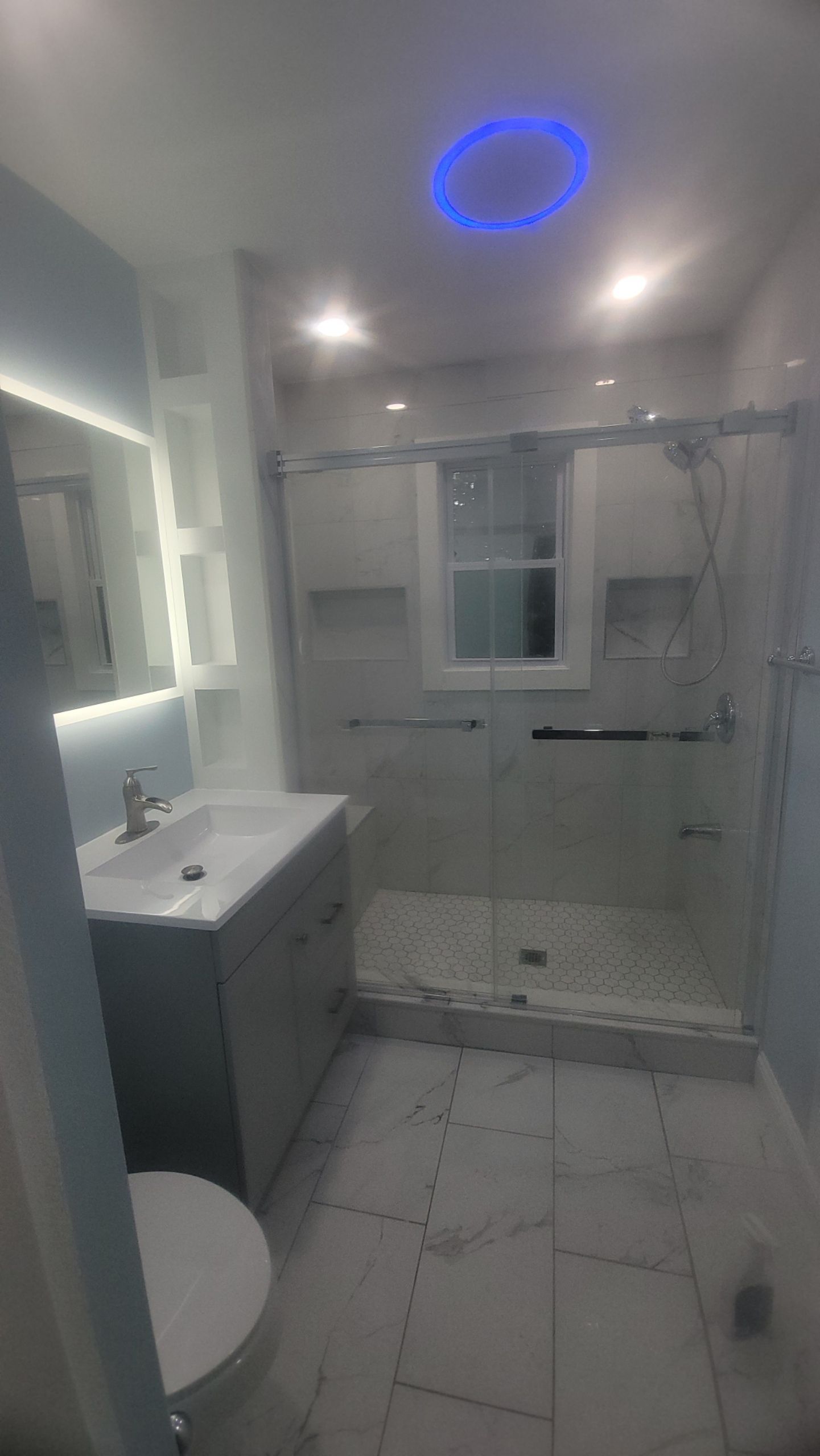Modern bathroom with gray vanity, glass shower, white and gray tile, and blue accent lighting.