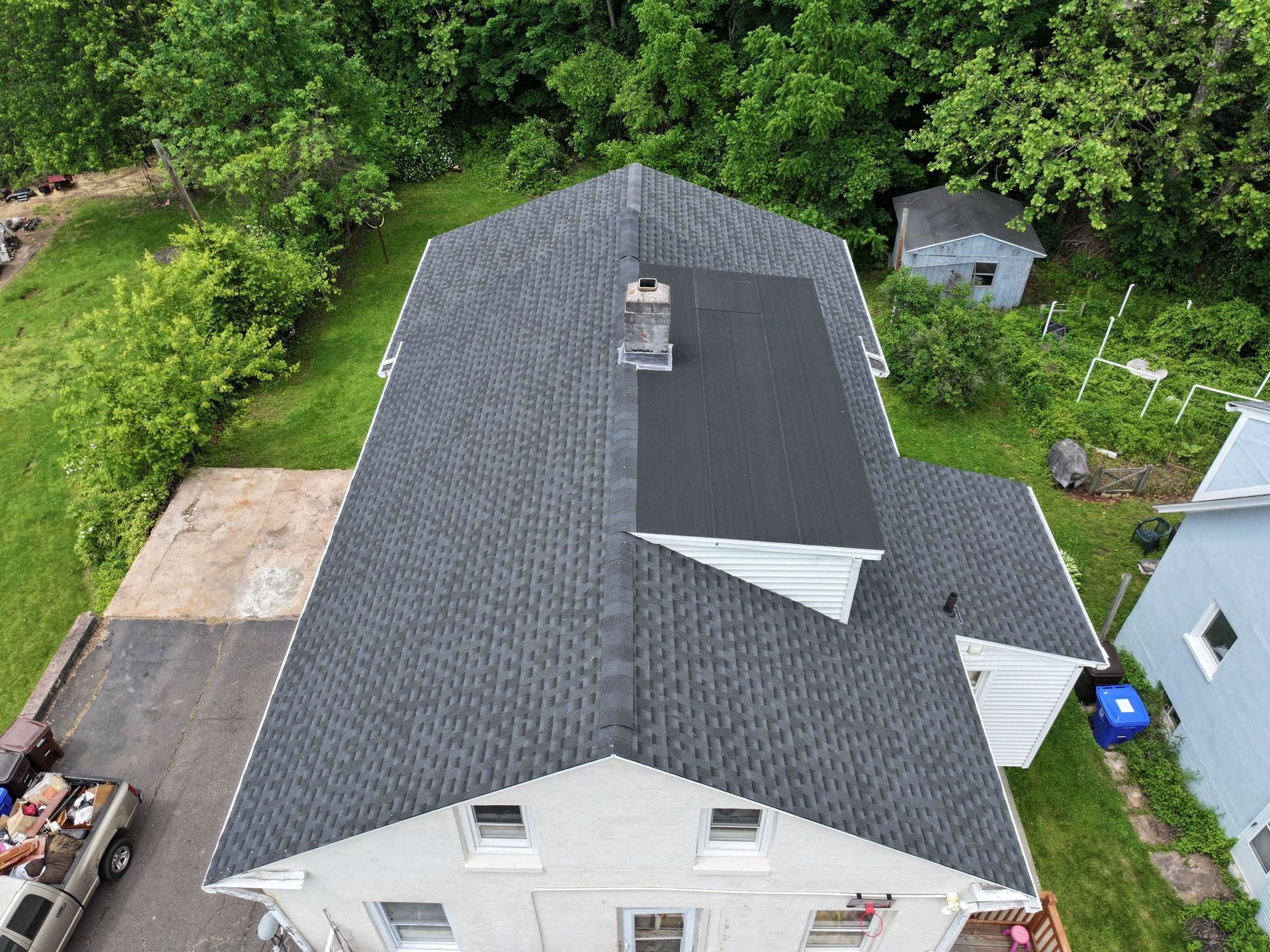 Overhead view of a house with a gray asphalt shingle roof, chimney, surrounded by trees and a driveway.