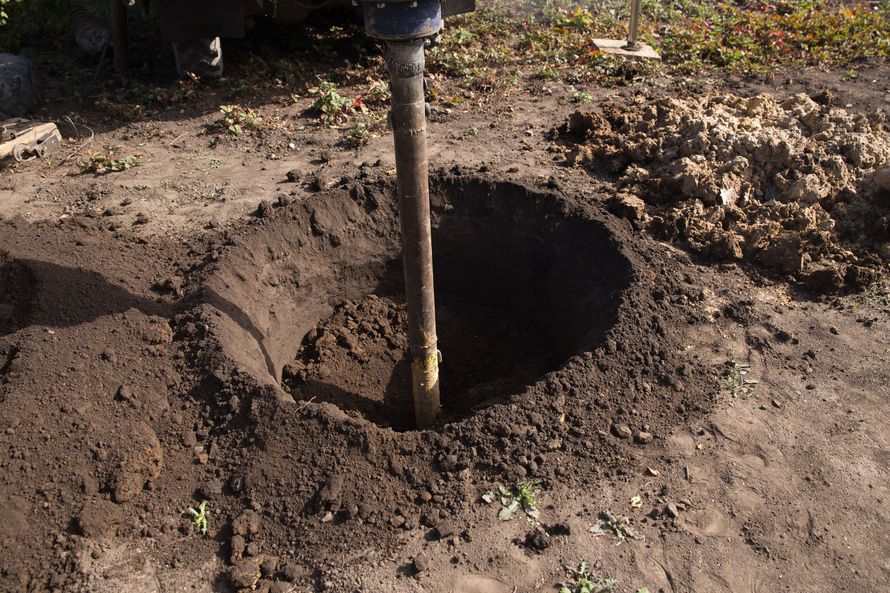 A metal pole is centered in a freshly dug circular hole in the dirt, likely for a fence post installation.