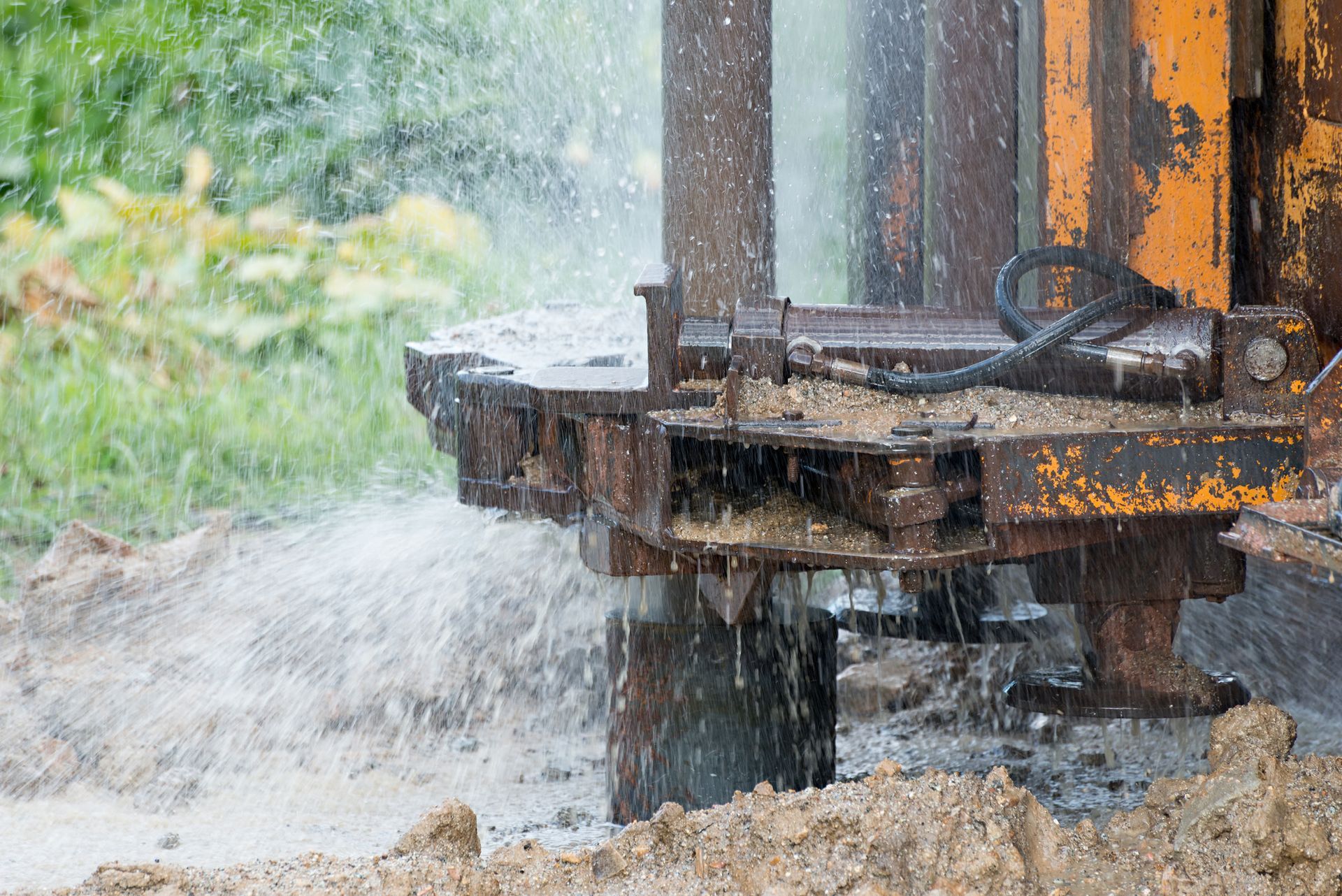 Close-up of a yellow industrial drill rig boring into muddy ground, spraying water and debris during a drilling operation.