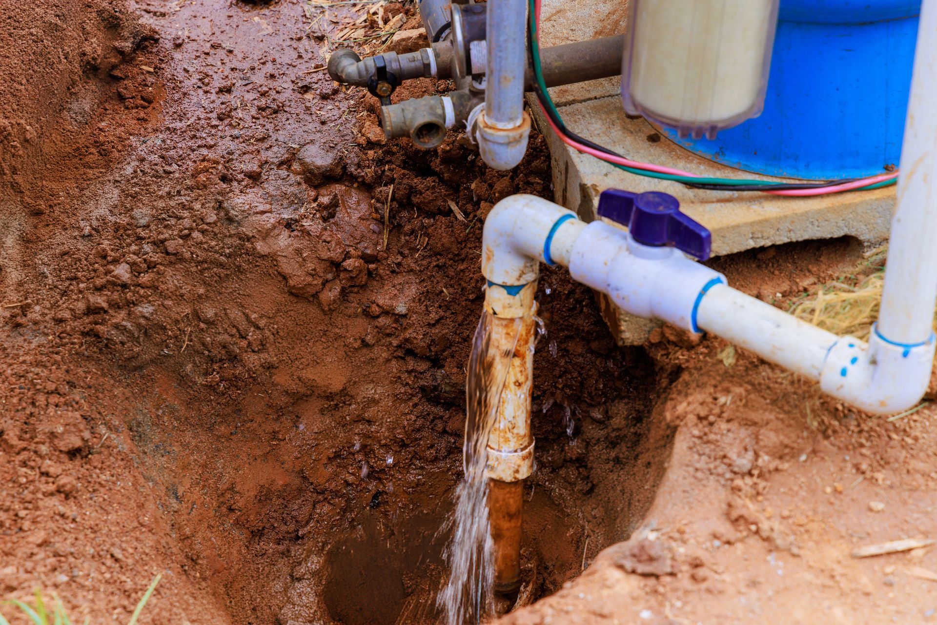 A leaky PVC pipe joint sprays water into a dirt trench next to a pump and filtration system.