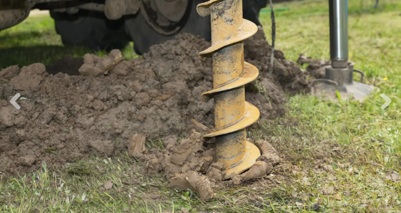 A yellow industrial auger drills into soil on a grassy lawn, with a mound of dirt beside it.