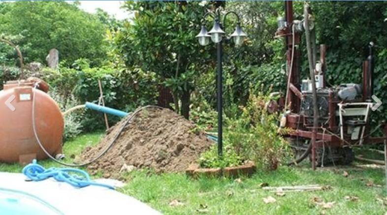 A small drilling rig operates in a backyard next to a pile of excavated dirt and a large brown water tank.