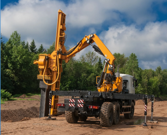 Yellow truck-mounted pile driver machine operating on a construction site with a forest background under a cloudy sky.