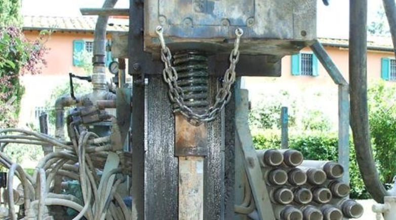 Industrial drilling rig mechanism with hydraulic hoses and stacked metal pipes, set in an outdoor, residential area.