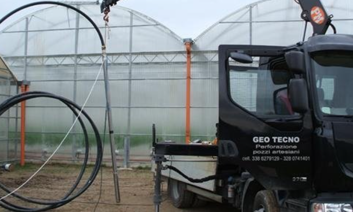 A black GEO TECNO service truck with a crane parked outdoors next to large greenhouse structures.