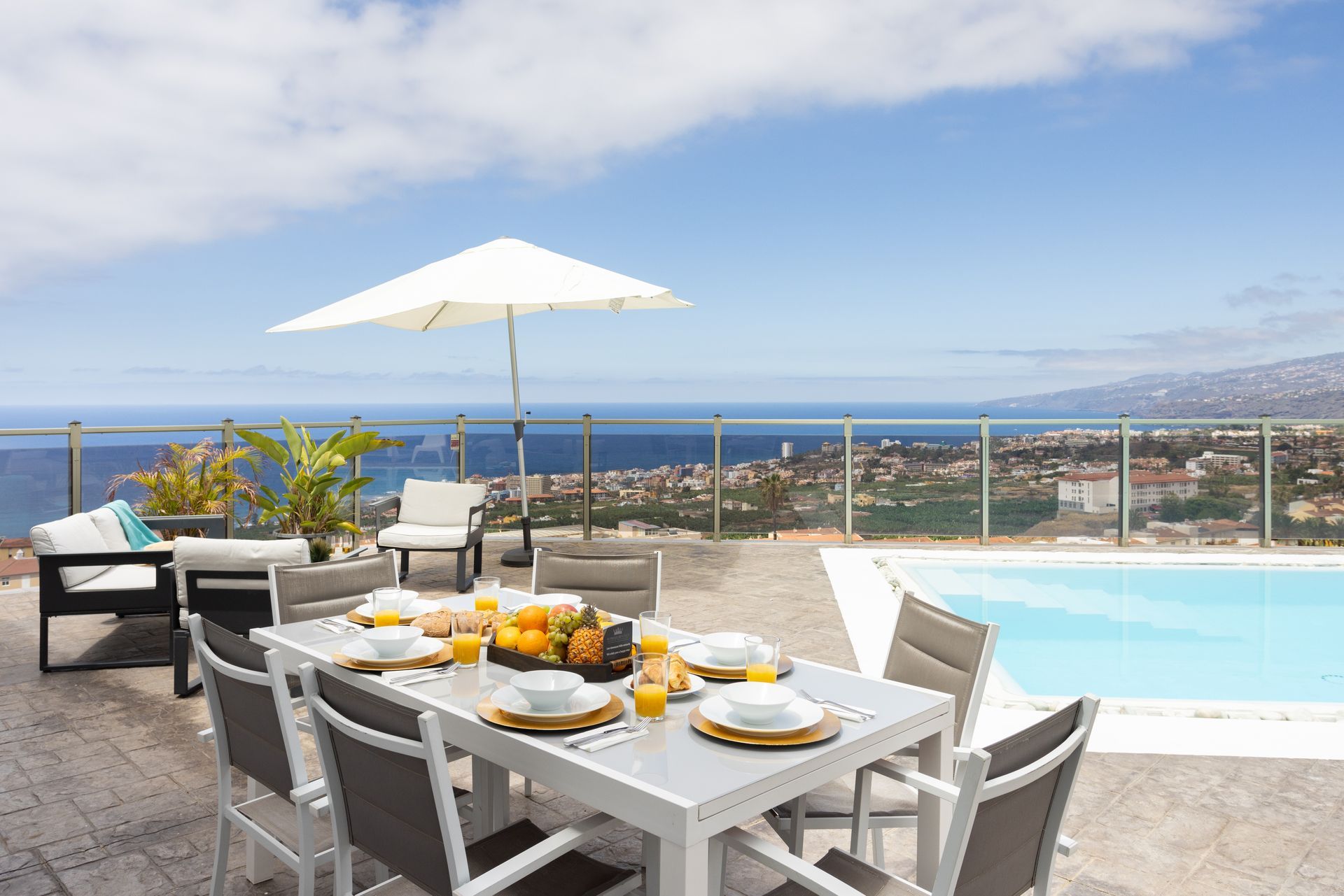 Mesa de comedor junto a la piscina con desayuno, sombrilla blanca, tumbonas y vista al mar.